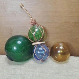 May include: Four decorative glass spheres in shades of green, blue, and gold. Two spheres are encased in netting, one with a jute string. The spheres vary in size and are arranged on a light-colored surface.