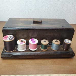 May include: A rectangular dark brown wooden sewing box with a lid and a small handle. The front base features a ledge holding six assorted spools of thread in various colors including black, white, pink, green, light blue, and dark brown.