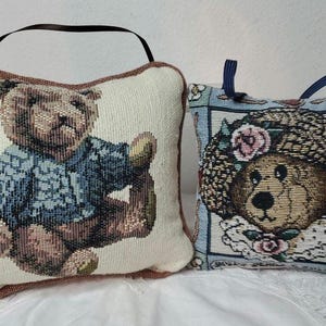 May include: Two embroidered throw pillows featuring teddy bears. One pillow is cream with a brown teddy bear wearing a blue sweater. The other pillow is blue with a brown teddy bear wearing a flower crown.