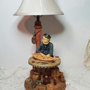 May include: A table lamp with a white lampshade and a nautical theme. The base features a figurine of a sailor working on a wooden boat, surrounded by nautical objects like barrels, rope, and tools.