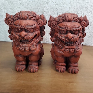 May include: A pair of terracotta-colored guardian lion statues, also known as shishi, featuring intricate carved details on the manes, faces, and paws. Each figure is depicted in a seated position with an open mouth, displaying teeth and a textured surface.