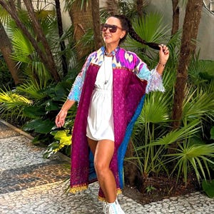 Puede incluir: Una mujer lleva un kimono largo de estampado rosa y azul con un vestido blanco debajo. Lleva zapatillas blancas de plataforma y tiene el pelo recogido en una coleta. Está de pie en un camino de piedra frente a un exuberante jardín verde.