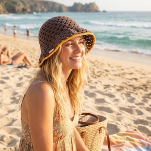 May include: A brown crochet bucket hat with a yellow brim is worn by a person on a beach. The hat has an open weave design. The person is wearing a floral print dress and is sitting on a colorful blanket. A woven tote bag sits nearby.