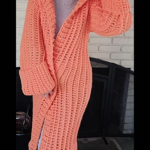 May include: A long, orange, hooded cardigan. This handmade, open-front sweater has long sleeves and a knee-length design. The chunky knit cardigan is a solid, vibrant orange colour.