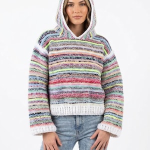 May include: A colorful, striped hoodie with a hood and long sleeves. The sweater features a mix of bright colors, including pink, yellow, and blue, with white trim on the hood and cuffs. The model is wearing blue jeans.