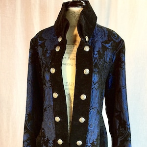 May include: A long, black and blue patterned coat with silver buttons. The coat features a high collar and velvet accents. The blue fabric has a floral design. The coat is displayed on a mannequin.
