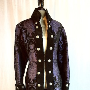 May include: A long, black and purple brocade coat with silver buttons. The coat features a high collar and a unique tailcoat design. The fabric has a detailed floral pattern. The coat is displayed on a mannequin.