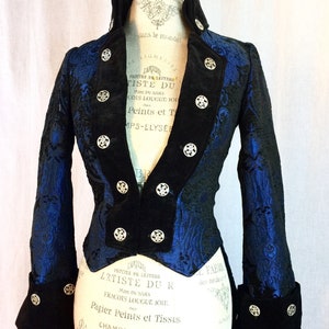 May include: A blue and black brocade tailcoat with silver buttons. The tailcoat has a velvet collar and cuffs. The fabric has a floral pattern. The tailcoat is on a white mannequin.