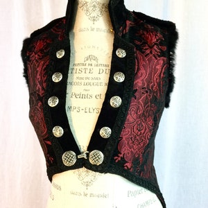 May include: A burgundy and black vest with ornate silver buttons and a decorative clasp. The vest features a high collar and is trimmed with black lace and faux fur. The fabric has a floral pattern.