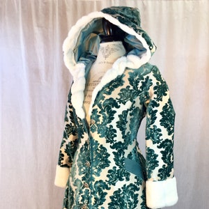May include: A green and white hooded coat with a damask pattern and white fur trim. The coat has a long, flowing skirt and a fitted bodice.