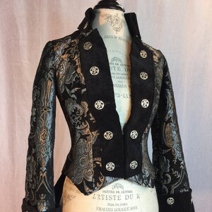 May include: A black and silver brocade jacket with a high collar and decorative silver buttons. The jacket features a tailored fit, with detailed floral patterns woven into the fabric. The cuffs also have silver buttons. The jacket is displayed on a mannequin.