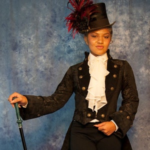 May include: A person wearing a black jacket with gold buttons, a white ruffled shirt, and black pants. They are holding a black walking stick and wearing a black top hat with a red and black feather.