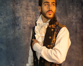 May include: A man wearing a white shirt with a ruffled collar and a blue and gold brocade vest. He is holding a wooden walking stick with a white handle.