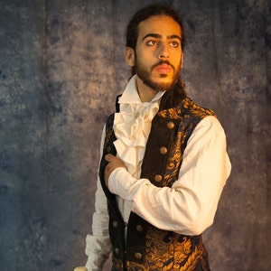 May include: A man wearing a white shirt with a ruffled collar and a blue and gold brocade vest. He is holding a wooden walking stick with a white handle.