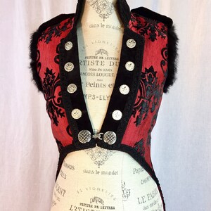 May include: A red and black sleeveless vest with a high collar and silver buttons. The vest has a black trim and a black floral pattern. The vest is on a mannequin with a white background.