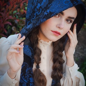May include: A woman with long brown braids wears a white blouse with a blue and black patterned hooded scarf. The scarf has a floral design.