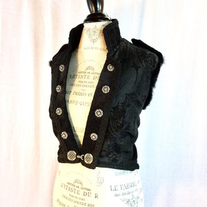 May include: A black sleeveless vest with a brocade pattern and silver buttons. The vest has a high collar and a fitted silhouette. The vest is decorated with silver buttons and trim. The vest is displayed on a white mannequin.