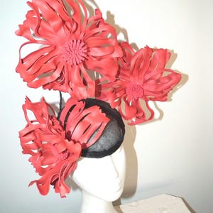 May include: A headpiece featuring three large, vibrant red flowers with long, curved petals and textured centers. The flowers are attached to a small, rounded black mesh base that sits atop a white mannequin head, creating a bold contrast.