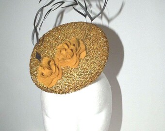 Puede incluir: Un sombrero fascinador redondo con una textura de purpurina dorada brillante. El tocado está adornado con dos flores de tela de color amarillo mostaza y una serie de plumas finas, curvas y de color gris oscuro que se extienden hacia arriba.