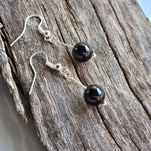 May include: A pair of dangle earrings featuring a polished black bead suspended from a silver-tone hook. The earrings are displayed on a weathered, textured wooden surface, showcasing their simple elegance and reflective quality.