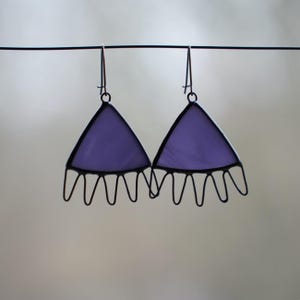 May include: A pair of lavender-colored triangle earrings with black metal detailing. The earrings have a unique design with a scalloped edge, suspended from thin black wires. The background is a soft, blurred neutral color.