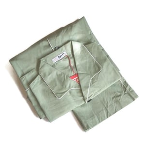 Könnte beinhalten: Salbeigrünes Pyjama-Set mit weißem Paspel. Das Set beinhaltet ein Oberteil mit Kragen und eine passende Hose, ordentlich gefaltet. Ein Etikett mit dem Markennamen ist am Oberteil befestigt. Der Pyjama ist aus einem weichen, leichten Stoff.