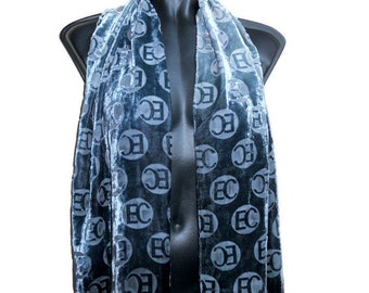 May include: A blue velvet scarf with a repeating pattern of the letters "EC" in white circles. The scarf is draped over a black mannequin, showcasing its texture and design. The scarf is a fashion accessory.