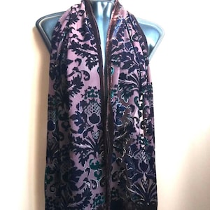 May include: A mauve scarf with a dark navy floral pattern and a brown velvet trim. The scarf is draped over a mannequin, showcasing its length and design. The floral pattern includes stylized leaves and other botanical elements.