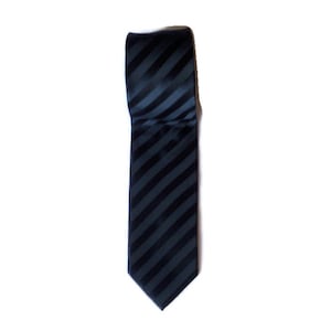 May include: A navy blue tie with dark blue diagonal stripes. The tie is folded over and lies on a white background. The tie is made of a smooth, shiny material.