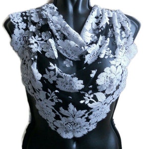 May include: A light gray lace scarf with a floral pattern. The scarf is draped over a black mannequin, showcasing the intricate details of the lace. The delicate design and color make it a versatile accessory.