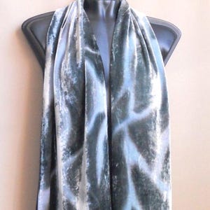 May include: A long scarf featuring a deep forest green and white tie-dye pattern. The fabric has a soft, velvet-like texture with a subtle sheen that catches the light, creating a flowing, draped appearance across the length of the garment.