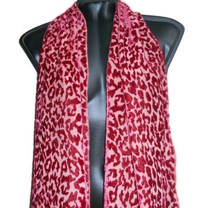 May include: A pink and burgundy scarf with a leopard print pattern. The scarf is draped over a black mannequin, showcasing its design. The fabric appears soft and lightweight, suitable for adding a touch of style.