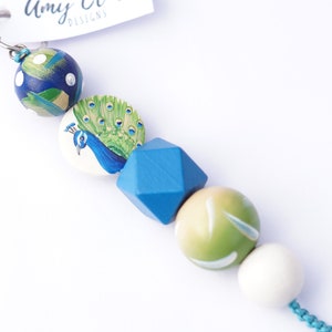 May include: A colourful wooden bead keychain featuring a peacock design. The beads are blue, green and white. The keychain is attached to a silver metal ring.