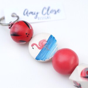 May include: A colourful wooden bead keyring with a red ladybug, a white bead with a pink flamingo on blue water, a red bead, a white bead with a red ladybug, and a black bead. The keyring is attached to a silver ring.