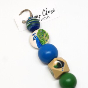 May include: A peacock themed keyring with blue, green and gold wooden beads. The beads are strung on a silver key ring. The keyring has a small tag that says "Tiny Close Designs".