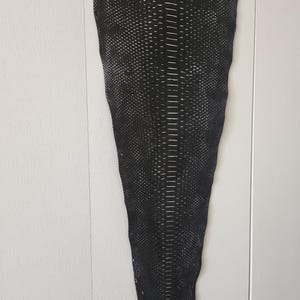 May include: A piece of black leather with a textured, scale-like pattern. The leather is rectangular with a tapered shape, and it hangs against a white wall. The pattern is detailed with small, raised scales.
