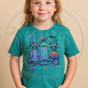 May include: A teal short-sleeve t-shirt featuring a graphic of two raccoons and a ghost in iridescent colors. One raccoon wears a witch hat, the other holds a pumpkin. The design includes a jack-o'-lantern, a spiderweb, and the text Spooky and Feral.