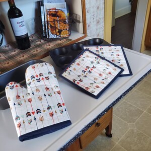 Peut inclure: Un gant de cuisine blanc et deux dessous de plat carrés assortis avec une bordure bleu marine. Le tissu présente un motif répété de flèches colorées. Un moule à muffins gris foncé et un moule à pain sont également visibles.