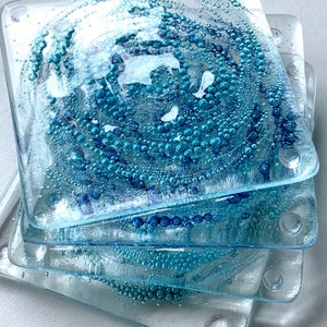 May include: Set of four blue glass coasters with a white swirl design and a bubble texture. The coasters are stacked on top of each other.