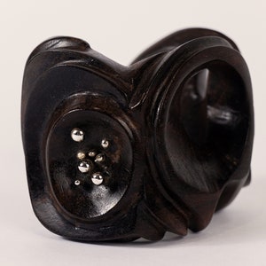 May include: A dark brown wooden bracelet with a unique, abstract design. The bracelet features a circular, recessed area with small silver dots.