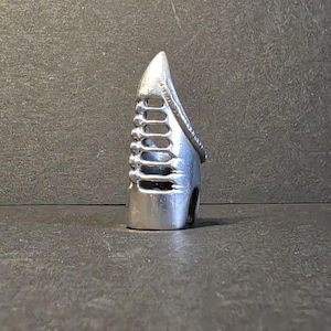 Könnte beinhalten: Ein silberfarbener Fingerpick mit spitzer Spitze und einer Reihe rechteckiger Aussparungen an der Seite. Der Pick hat ein gebogenes Design und ein kleines Loch in der Nähe des Bodens.