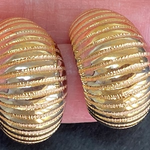 May include: A pair of gold-colored, textured earrings. Each earring has a curved, oval shape with horizontal ridges. The earrings appear to be made of metal and reflect light, suggesting a polished finish. The design is simple yet elegant.