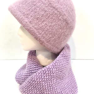 May include: A light purple knit hat and infinity scarf set. The hat has a rounded crown and a small brim. The scarf is a wide, knitted loop. Both items are made of a soft, textured yarn, perfect for cold weather.