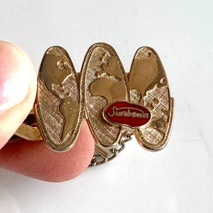 May include: Gold-tone tie clip with a chain and three globe-shaped charms. The center charm has a red enamel logo that says "Sunbeam".