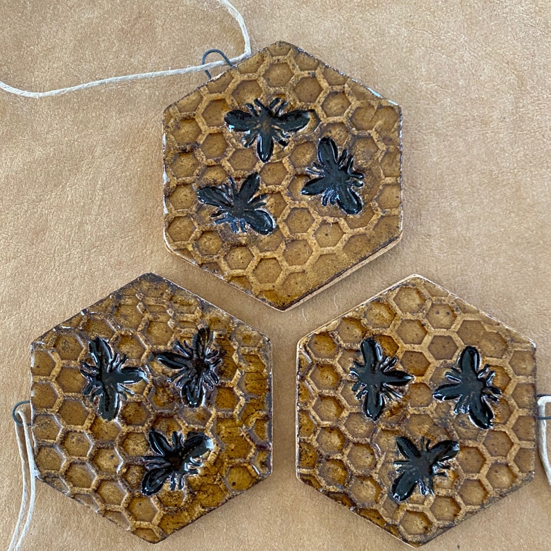 May include: Three hexagonal ceramic tiles with a honeycomb pattern and black bee silhouettes. The tiles are brown and gold in colour.