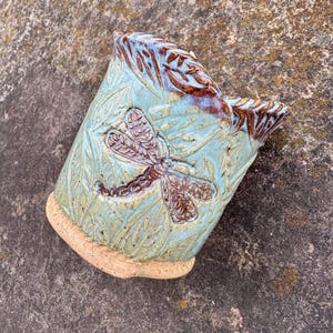 May include: A handmade ceramic planter with a dragonfly design. The planter is a light blue color with brown accents and a textured surface. The dragonfly is brown and is the focal point of the design. The planter has a unique shape and is suitable for small plants.