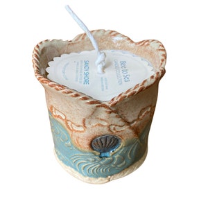 May include: A ceramic candle holder with a blue and brown glaze. The holder features a wave design and a seashell. The candle is labelled "Bee to Sea Candle Collection" and "Sandy Shore with calming notes of sarilla and citrus".