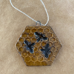 May include: A hexagonal ceramic ornament with a honeycomb pattern and three black bees. The ornament is brown and has a string attached for hanging.