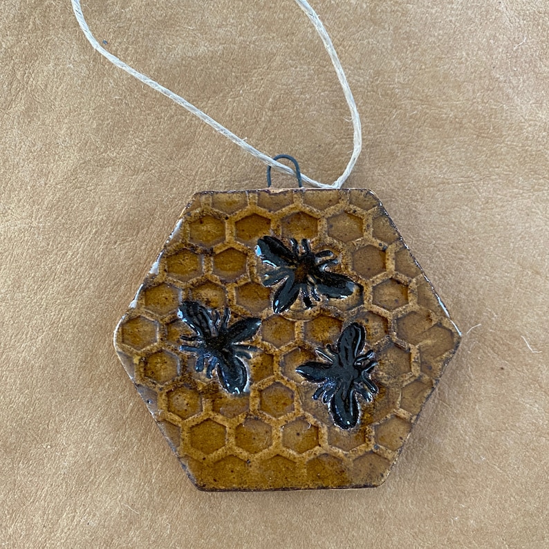 May include: A hexagonal ceramic ornament with a honeycomb pattern and three black bees. The ornament is brown and has a string attached for hanging.