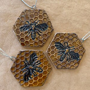 May include: Three hexagonal ceramic ornaments with a honeycomb pattern and a black bee silhouette on each. The ornaments are strung with twine.